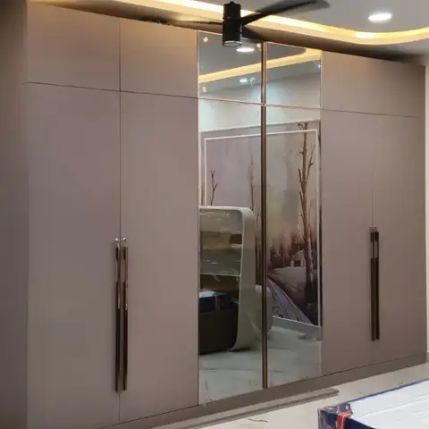 Bedroom with a mauve wardrobe wall with mirrored centre shutters and gold bar handles, new furniture still under protective plastic wrap in the foreground
