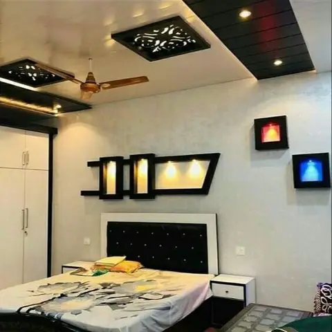 Bedroom with a multi-tier POP false ceiling featuring carved jaali cutouts above a black tufted headboard, flanked by red and blue lit wall display boxes.