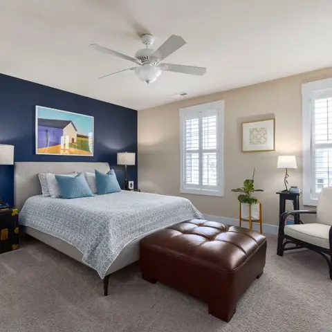 Bedroom with a navy accent wall behind an upholstered bed, a leather tufted bench at the foot, and a rattan armchair with a small side table by the window.
