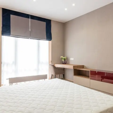 Bedroom with a navy-trimmed roman blind at the window, a built-in study desk, and a floating console in beige and glossy maroon panels beside a bare mattress