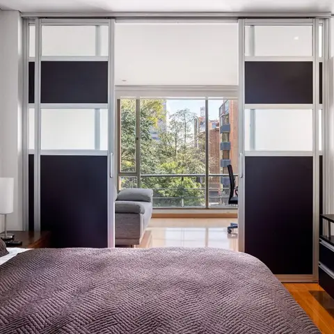Bedroom with a purple quilted bedspread facing sliding partition doors that open to an adjoining study nook with a desk chair and city view