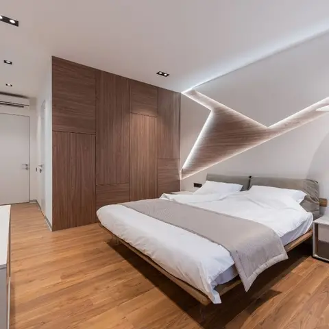 Bedroom with a sculptural angular cove-lit ceiling fin in wood veneer above the bed, a full-height wood-shutter wardrobe wall