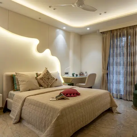 Bedroom with a sculptural backlit wave-shaped headboard wall, cove-lit ceiling, and a green bouclé lounge chair beside the window