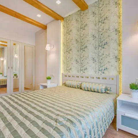 Bedroom with a slatted white headboard against a backlit blossom-print wallpaper panel, a striped sage bedspread with a matching bolster