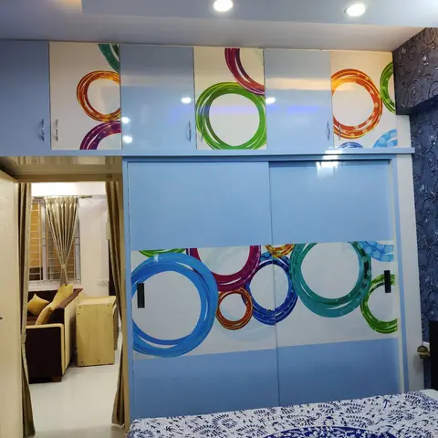 Bedroom with a sliding wardrobe featuring a colourful ring-pattern print and dark blue floral wallpaper, glimpsed through to the living room beyond.