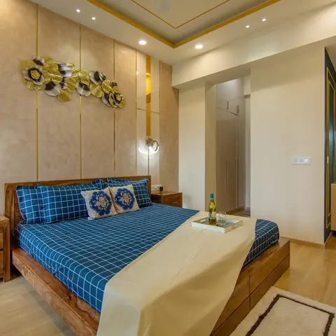 Bedroom with a solid wood bed in blue check linen, a gold metal floral wall sculpture on a brass-inlaid panel wall, and a glass-doored walk-in wardrobe beyond.