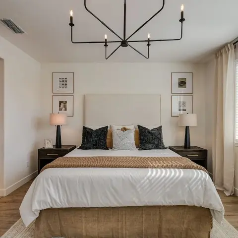 Bedroom with a tall linen headboard flanked by symmetrical nightstands and a black candle-style chandelier, opening directly onto an ensuite bathroom.
