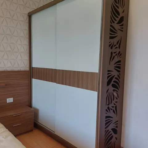 Bedroom with a tall two-tone sliding-door wardrobe featuring a carved leaf-motif jaali side panel, set against a geometric-textured wallpaper wall