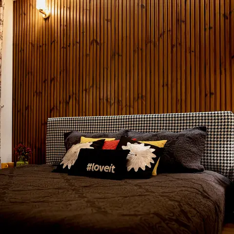 Bedroom with a vertical wood-slat panelled wall behind a houndstooth upholstered headboard, dark quilted bedspread with a graphic '#loveit' cushion