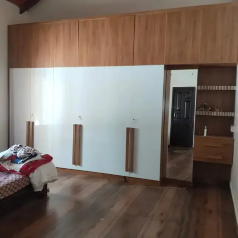 Bedroom with a wall-to-wall wardrobe featuring wood-finish loft units above glossy white lower shutters with wood pull handles