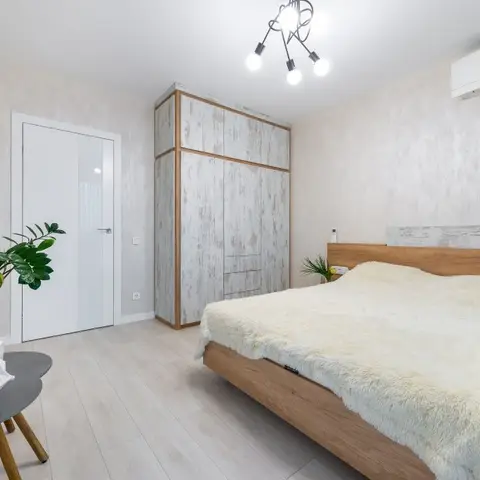 Bedroom with a whitewashed distressed-finish wardrobe, wood-trimmed bed frame topped with a cream faux-fur throw, and textured wallpaper