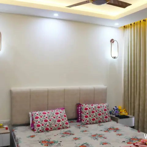 Bedroom with a wide beige channel-tufted headboard flanked by two oval brass-and-glass wall sconces, floral bedding and side tables holding a child's toys