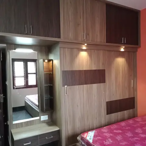 Bedroom with a wood-laminate wardrobe wall, an integrated dressing mirror unit, and a bed with its mattress still in plastic wrap.