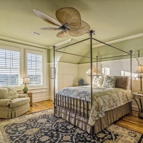 Bedroom with a wrought-iron canopy bed, sage-green walls above white wainscoting, and a tropical palm-blade ceiling fan.