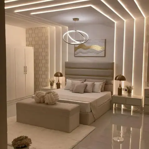 Bedroom with an elaborate cove-lit stepped ceiling outlining a ring pendant chandelier, a channel-tufted headboard with brass trim
