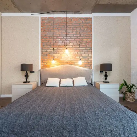 Bedroom with an exposed brick accent wall behind the headboard, three bare-bulb pendant lights hanging from a raw concrete ceiling