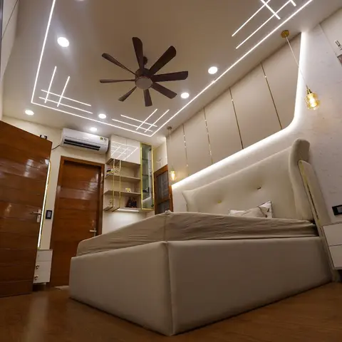 Bedroom with an ornate multi-tier cove-lit false ceiling in a linear pattern, a wood-bladed ceiling fan, a wingback tufted cream headboard trimmed in gold