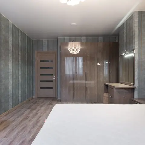 Bedroom with damask-patterned wallpaper, a dark wood mirrored wardrobe, and a matching lit vanity dresser with wall sconces beside the bed.