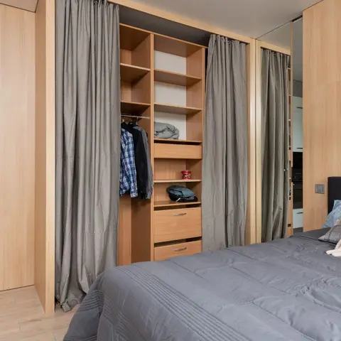 Bedroom with full wood-panelled walls and an open closet system dressed with grey curtain doors, dark upholstered headboard, and a plush toy on the pillow.