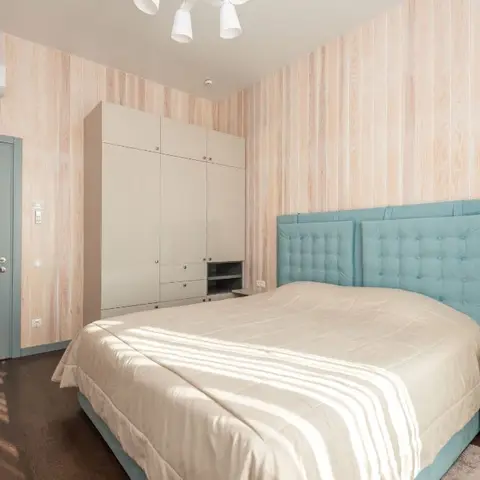 Bedroom with light wood-panel wallpaper, a teal tufted upholstered headboard, a full-height white wardrobe wall, and a sage-green painted door.