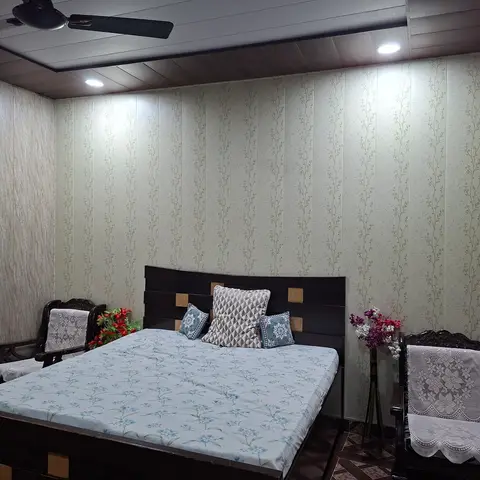 Bedroom with pale floral wallpaper panels, a dark wood-paneled cove ceiling, and a wooden bed dressed in a blue floral bedspread flanked by carved wooden
