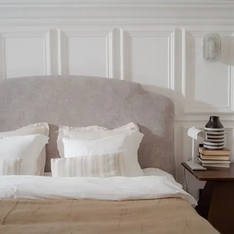 Bedroom with white raised-panel wainscoting behind a curved linen-upholstered headboard, layered white and tan bedding