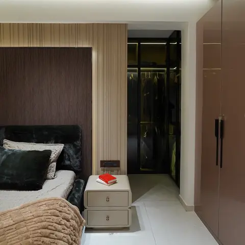 Bedside view of a dark walnut headboard wall beside a fluted-panel doorway leading into a glass-front walk-in wardrobe.