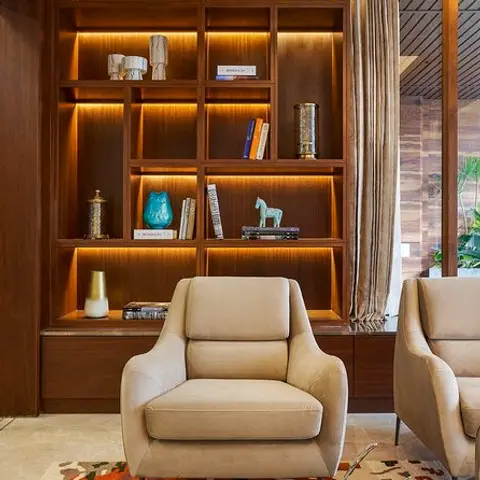 Beige armchairs in front of a backlit wooden display shelving unit styled with books and objets, with a bold multicolour patterned rug underfoot.