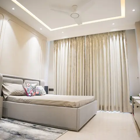 Beige bedroom with a raised-panel moulded headboard wall, box cove-lit false ceiling, floor-length curtains and a wall-mounted TV over a low console