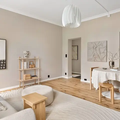 Beige living-dining room with a cream sofa and pouf in the foreground, a round wood dining table under a sculptural white pendant light