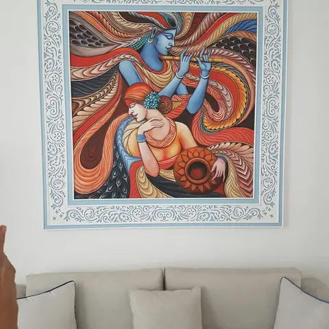 Beige sofa with scatter cushions below a large framed traditional Radha-Krishna painting in an ornate light-blue border.
