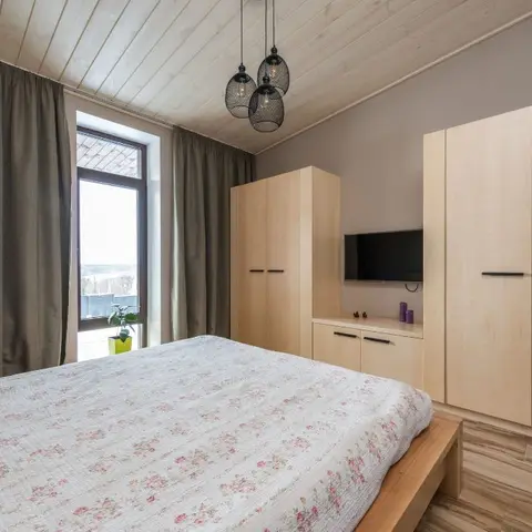 Birch-faced wardrobes and a low TV cabinet under a white-washed plank ceiling, with olive-grey floor curtains and a black mesh cluster pendant over a