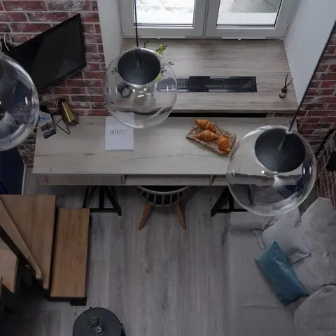 Bird's-eye view of an industrial loft mezzanine with exposed red-brick walls, glass globe pendant lights over a light-wood desk, a wooden staircase