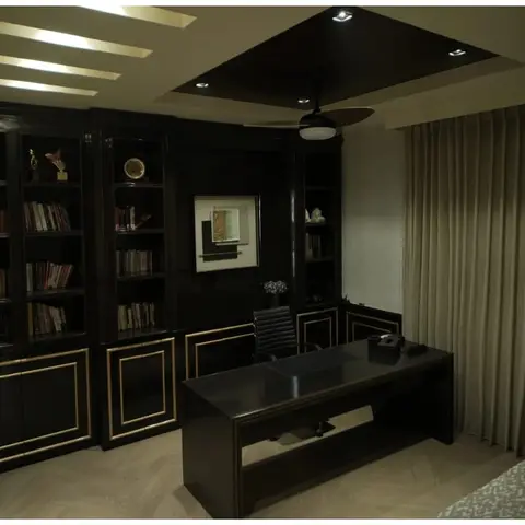 Black lacquered built-in bookshelf and gold-trimmed executive desk in a cove-lit home study, with a herringbone wood floor and beige curtains.