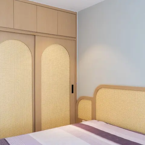 Blond-oak sliding wardrobe with arched natural cane-webbing inserts and a loft above, matched by a cane-panel headboard against a pale blue wall.
