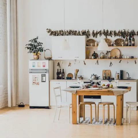 Boho dining nook with a rustic wood table and mismatched chairs, dried herb garlands strung above open kitchen shelving against a whitewashed brick wall.