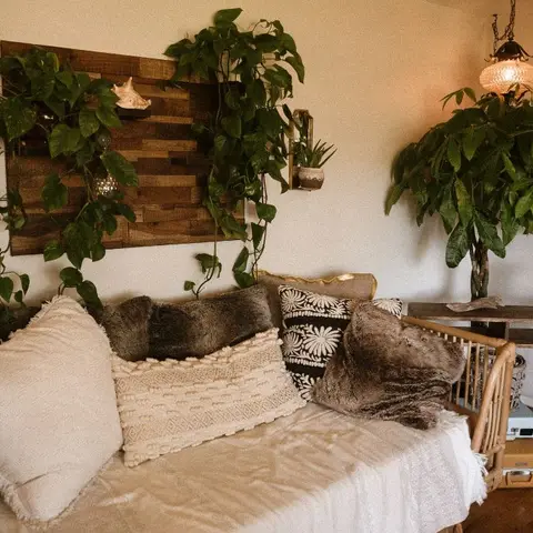 Boho living room with a cushion-piled cream sofa beneath trailing potted plants and a reclaimed-wood wall art piece, lit by a vintage glass pendant lamp