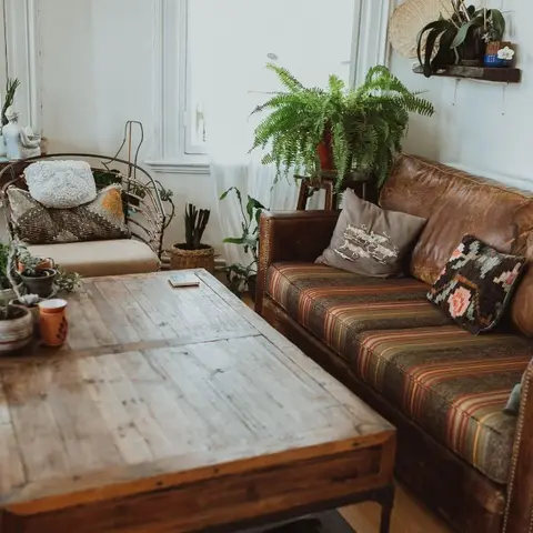 Boho living room with a tan leather sofa layered with kilim-pattern cushions, a reclaimed wood coffee table, and abundant potted plants by the window.