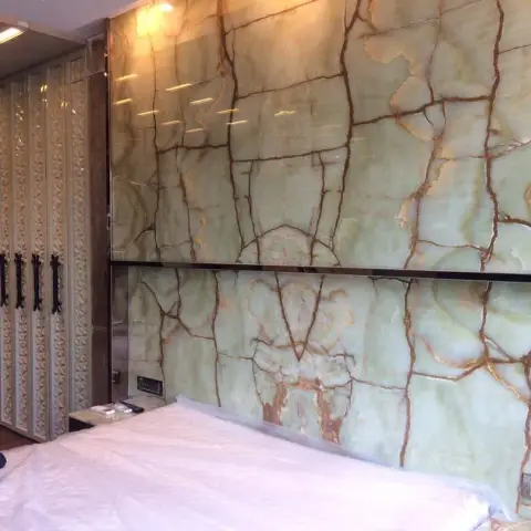 Book-matched green onyx slab headboard wall with a dark reveal band splitting it in two, facing a run of champagne CNC-carved wardrobe shutters