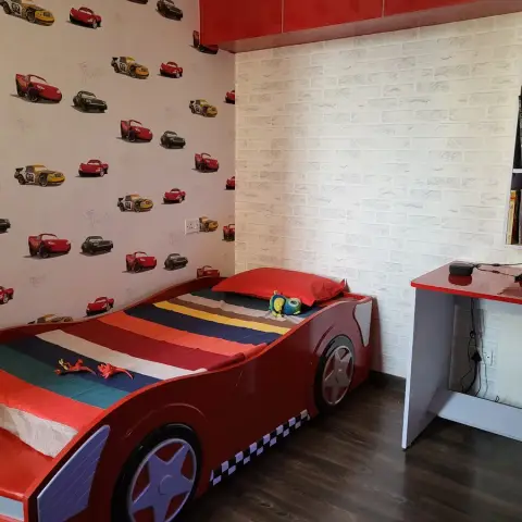 Boy's room with a red racing-car bed made up in a striped blanket, a cartoon-car print wallpaper on one wall, white brick-effect wallpaper on the other