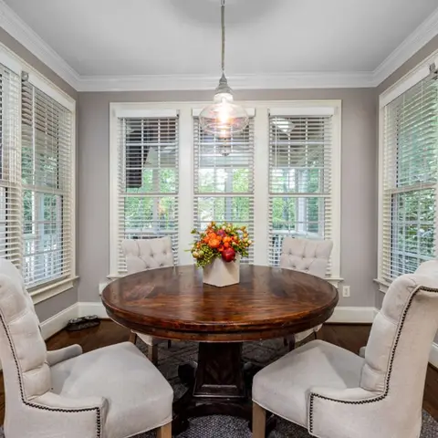 Bright breakfast nook wrapped in windows with white blinds, a round dark-wood pedestal table and cream tufted wingback dining chairs under a single glass