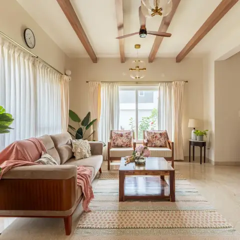 Bright living room crossed by teak ceiling beams with a wooden-blade fan and brass globe pendants, a teak-framed sofa under a coral throw