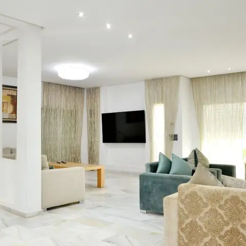 Bright living room with a wall-mounted TV, teal and gold damask-pattern sofas, polished marble flooring and floor-to-ceiling sheer curtains opening onto a