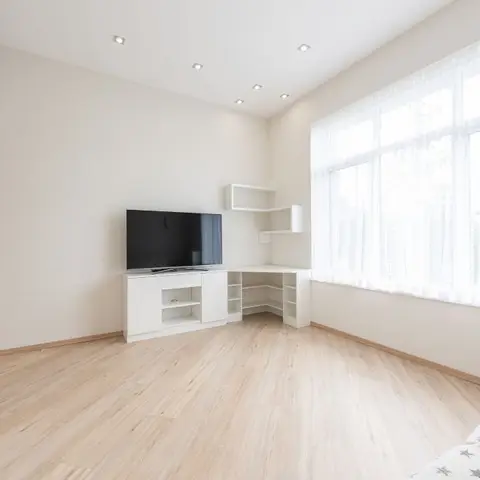 Bright minimal room with a white corner TV unit with open display shelving, light wood flooring, and sheer white curtains across a large window.