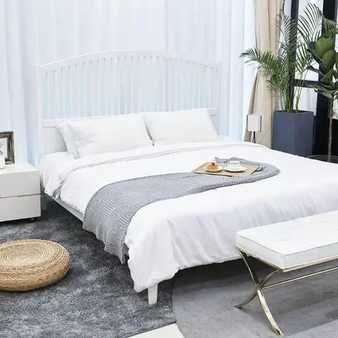 Bright white bedroom with a spindle-slat headboard bed, grey diamond-weave throw, jute pouf on a grey rug, and a gold-frame bench at the foot of the bed.