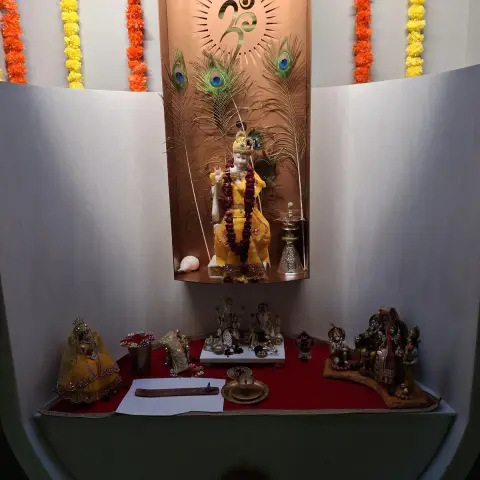 Built-in curved pooja niche with a backlit copper Om panel, peacock feathers and marigold garlands over a red-cloth altar shelf holding family deities.