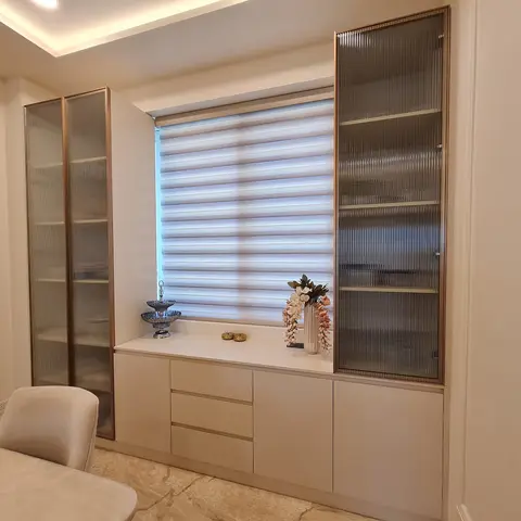 Built-in display cabinet with fluted glass doors and brushed-brass trim flanking a window with layered blinds, above a cream storage console with drawers.