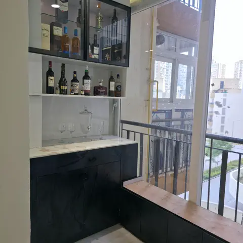 Built-in home bar unit with glass-front upper cabinet displaying liquor bottles and black cabinetry with marble counter beside a balcony window