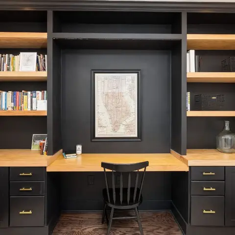 Built-in home office nook with black cabinetry and open wood shelving flanking a wood desk, a framed vintage map centered above, and a black spindle-back chair.