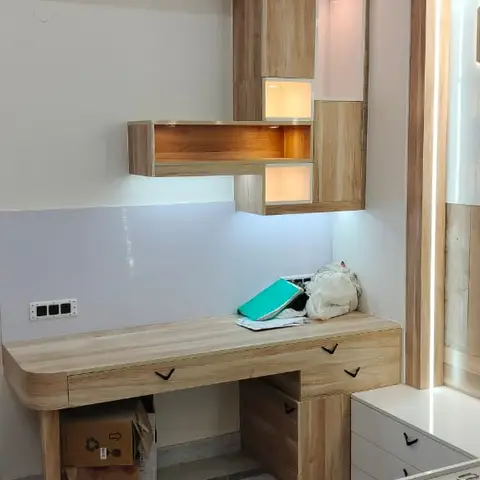 Built-in light-oak laminate study desk with a floating wall shelf unit and backlit display niches above, lit by ceiling cove lighting